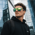 Mann mit grüner Sonnenbrille und schwarzem Kapuzenpullover, vor einem modernen Glasgebäude stehend.