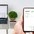 Hand hält Smartphone mit digitaler Rechnung "INVOICE PAID", Laptop und Pflanze im Hintergrund.