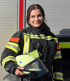 Eine Feuerwehrfrau in Einsatzkleidung hält einen hellgelben Helm mit Visier und lächelt in die Kamera.