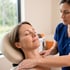 A therapist in blue scrubs performs a manual shoulder massage on a female patient resting in a treatment chair.