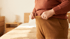 A person in a brown sweater pulls the waistband of their tan trousers away from their stomach in a bedroom.