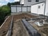 Gartenbaustelle mit Erdaushub, Randsteinen aus dunklem Beton und zwei Schubkarren vor einem weißen Wohnhaus.