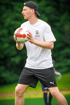Ein Mann in einem weißen T-Shirt mit „BUFFON FOOTBALL ACADEMY“-Logo und schwarzer Kappe hält einen Fußball.