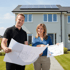 Lachendes Paar hält Baupläne und Diagramme vor einem Haus mit Solarmodulen.