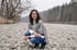 Frau mit Brille und grauem Poncho kniet an einem steinigen Flussufer vor bewaldeter Kulisse.