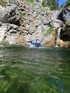 Eine Person in blauer Ausrüstung und Helm taucht aus grünem Wasser vor Felsen und Wasserfällen auf.