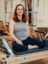 Lächelnde Frau mit roten Haaren sitzt im Schneidersitz auf einem Pilates-Reformer in einem Studio.