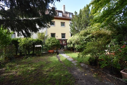 Hinterhof mit gelbem Mehrfamilienhaus, grünem Garten, Büschen, Bäumen und einem Steinweg.
