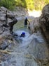 Zwei Personen in Neoprenanzügen und blauen Helmen klettern in einem felsigen Flussbett.
