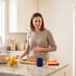 A woman in a kitchen grimaces while holding her stomach near a counter with chopped vegetables and lemons.