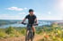 A man in a helmet and gear rides a mountain bike on a dirt trail overlooking a river valley and bridge.
