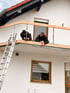 Zwei Handwerker in dunkler Arbeitskleidung montieren ein Holzgeländer an einem Balkon eines weißen Wohnhauses.