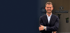 Lukas Tschanter von Finanzkonzepte lächelt im Anzug vor einem Logo an einer Holzlamellenwand.