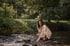 Frau in einem geblümten Kleid hockt am Ufer eines flachen Bachs im Wald und berührt das Wasser.