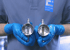 Hände in blauen Handschuhen halten zwei metallische Drosselklappen mit sichtbaren Lamellen.