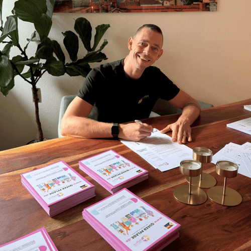 Ein lächelnder Mann in Schwarz unterschreibt Dokumente an einem Holztisch mit rosa Broschüren "QUEER PFLEGE".