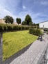 Gepflegter Garten mit grünem Rasen, Pflastersteinen, einer Hecke und einer Schlauchtrommel unter blauem Himmel.
