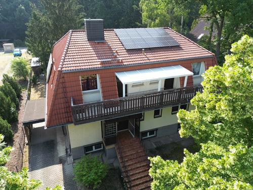 Haus mit rotem Ziegeldach, Solarpaneelen und einem Balkon, umgeben von grünen Bäumen.