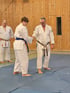 Zwei Männer in weißen Karate-Anzügen schütteln sich die Hände, ein dritter Mann steht im Hintergrund.