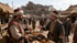 Two men in traditional Ottoman clothing and turbans talk at a crowded marketplace in front of a stone fortress.