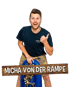 Micha von der Rampe lächelt in Lederhosen neben einer blauen Tuba vor schwarzem Hintergrund.