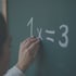 Eine Hand zeigt auf die mit Kreide geschriebene mathematische Gleichung 1 x = 3 auf einer grünen Schultafel.