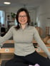 Lächelnde Frau mit Brille und grauer Sportjacke sitzt in einem Pilates-Studio vor Trainingsgeräten.