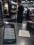 Smartphone mit Benachrichtigungen und Notizzettel „Rückruf Firma Meier“ in einer Werkstatt mit Industriearbeitern.