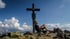 Ein Mann lehnt entspannt am Gipfelkreuz auf einem felsigen Berggipfel unter blauem Himmel mit Wolken.
