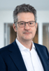 Ein Mann mit grauen Haaren und Brille trägt ein weißes Hemd und einen dunkelblauen Anzug.