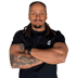 Muskulöser Mann mit Dreadlocks, Bart und Tattoos, der die Arme vor einer grünen Wand verschränkt.