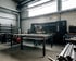 Ein Metallbauer mit Schweißmaske bearbeitet Metallteile an einem Werktisch in einer Industriehalle mit Funkenflug.