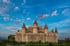 Ein sandfarbenes Steinschloss mit mehreren dunklen, spitzen Türmen unter einem blauen Himmel mit weißen Wolken.