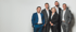 Fünf Geschäftsleute, drei Männer und zwei Frauen, lächeln in Anzügen vor weißem Hintergrund.