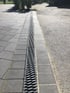 Lange Entwässerungsrinne mit schwarzem Metallgitter zwischen gepflastertem Gehweg und asphaltierter Straße.