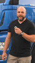 Ein bärtiger Mann mit schwarzem T-Shirt und beiger Hose hält ein Mikrofon vor einem blauen LKW.