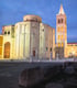 Eine beleuchtete Kirche mit Kuppel und ein hoher Glockenturm bei Dämmerung, umgeben von Steinsäulen und Ruinen,die Stadt nennt sich Zadar.