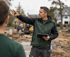 Ein Bauleiter im grünen Anorak zeigt auf einer Baustelle mit einem Tablet in der Hand.