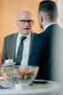 Ein Mann im Anzug hält eine Kaffeetasse und spricht mit einer anderen Person.