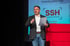 Ein Mann steht auf einer Bühne, hält Papiere und gestikuliert, vor einem Bildschirm mit dem SSH-Logo und Text "Arbeitssicherheit".