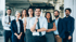 Ein lächelndes, diverses Team in Business-Kleidung steht gemeinsam in einem modernen, hellen Bürogebäude.