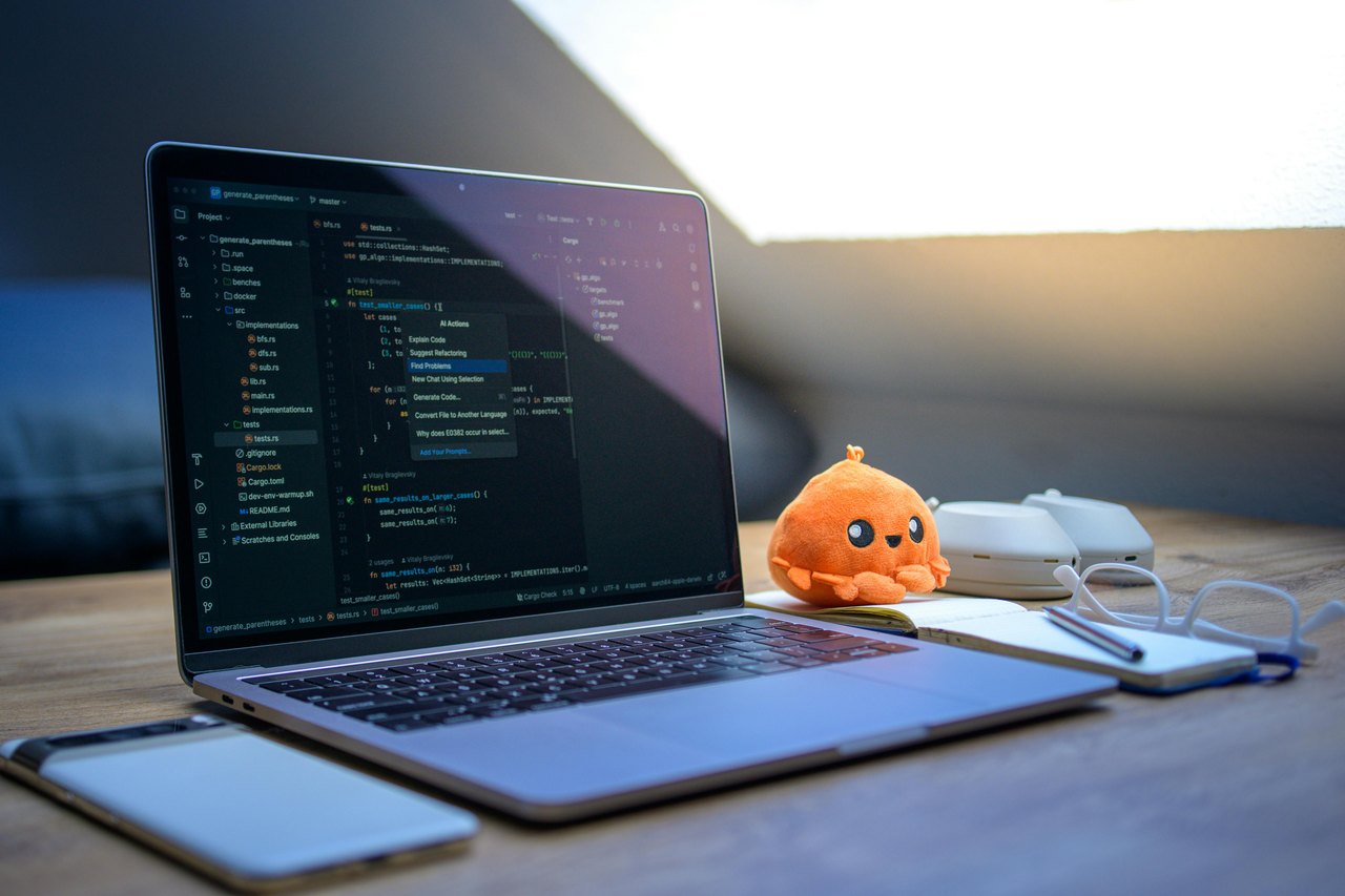 Laptop mit Code-Editor, Smartphone, Notizbuch und orangefarbener Oktopus-Plüschfigur auf einem Holztisch.