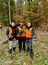 Drei Männer in orangefarbenen Westen stehen mit Schaufeln und jungen Bäumen im Wald.
