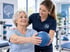 Physiotherapeutin unterstützt eine Seniorin bei einer Übung mit einem blauen Medizinball in einer Praxis.