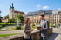 Ein Mann im Anzug spricht mit einer Frau vor einem Schloss, einem Brunnen und einer Wegweiser-Tafel.