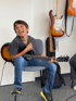A young boy with dark hair, wearing a gray shirt and jeans, sits on a chair playing an electric guitar. He laughs joyfully.