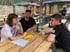 Drei Personen sitzen an einem Holztisch im Freien, diskutieren und lächeln; Notizen liegen vor ihnen.