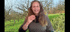 Eine lächelnde Frau mit langen Haaren und grauer Strickjacke gestikuliert vor einem virtuellen Hintergrund mit Bäumen und Wiese.