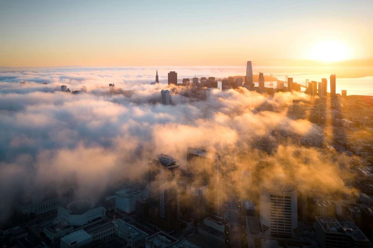 Luftaufnahme der Skyline von San Francisco bei Sonnenaufgang, wobei dichte Nebelschwaden zwischen den Wolkenkratzern ziehen.