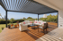 Moderne Pergola mit Lamellendach über einer Holzterrasse mit Lounge-Möbeln und Pflanzen.